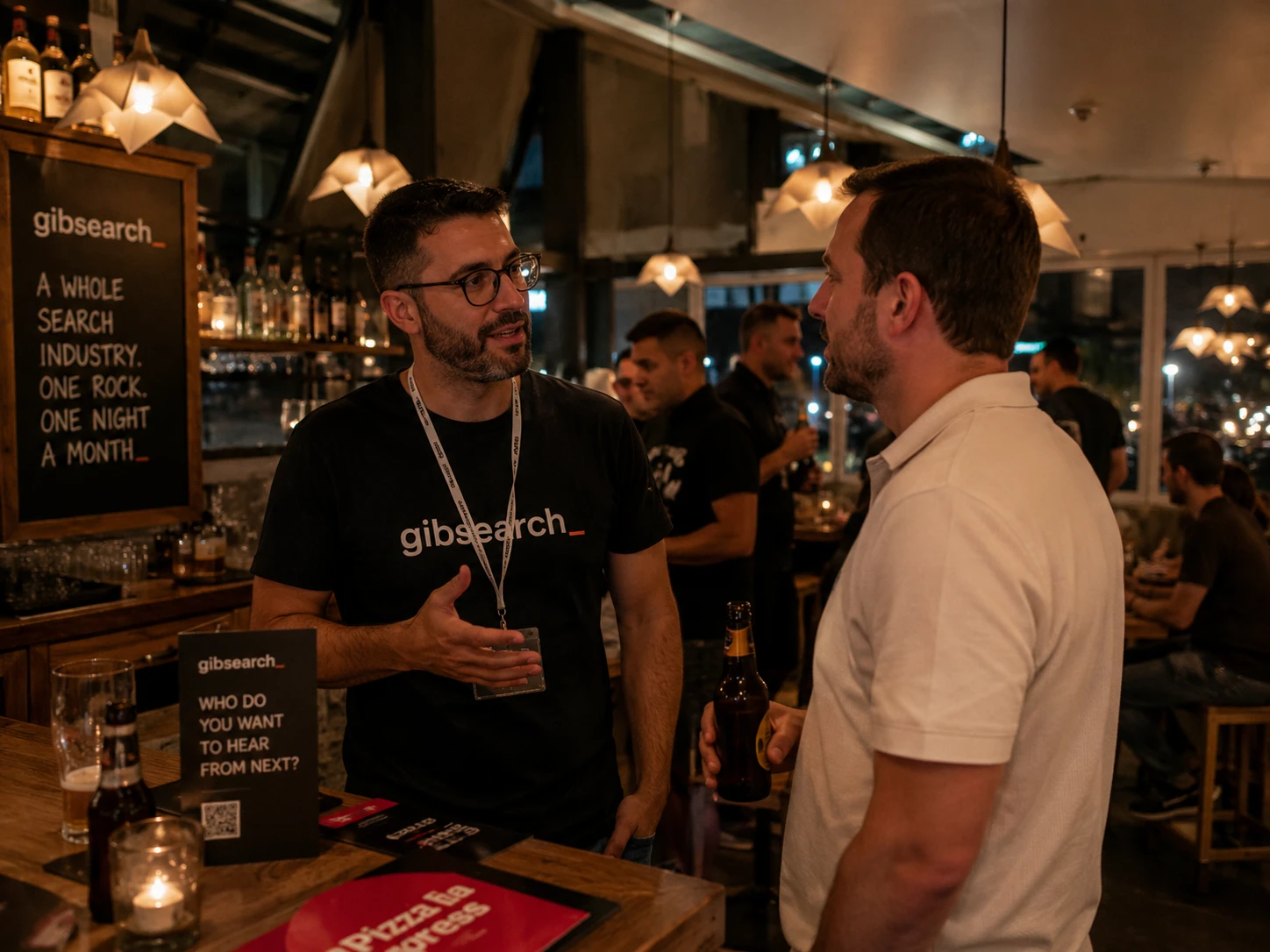 Two people talking at the bar at a GibSearch night, one wearing a GibSearch T-shirt, beer in hand.